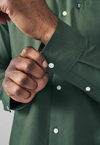 Camisa verde de botón con un tejido texturizado. Botones blancos en los puños. Mano visible ajustando el puño. Vista en primer plano.