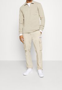Homme portant une chemise beige à manches longues avec fermeture quart de zip, un pantalon cargo beige avec poches, et des baskets blanches, sur fond blanc.