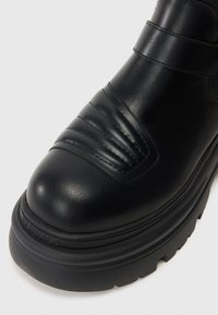 Μαύρη δερμάτινη μπότα στον αστράγαλο με ριχτές λεπτομέρειες και χοντρή, υφασμένη σόλα. Διαθέτει λείο φινίρισμα και έντονα ραφές.