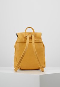 Hořčičně žlutý kožený batoh s nastavitelnými popruhy a horním madlem, stojící vzpřímeně na bílé ploše na pozadí bez vzoru.