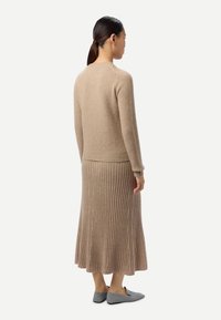 Femme aux cheveux foncés attachés porte un pull en maille beige et une jupe plissée assortie en maille, avec des chaussures grises sans lacets, debout de profil.