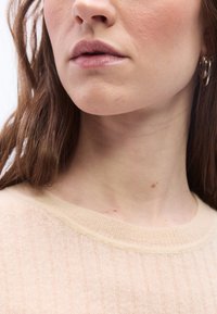 Gros plan sur le bas du visage et le cou d'une femme avec un maquillage léger, une boucle d'oreille en or et un pull côtelé beige.