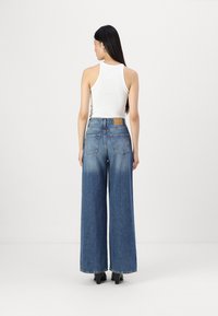 Vaqueros de pierna ancha en denim azul con cintura alta, con un parche de cuero en la parte posterior y dos bolsillos traseros. Acompañados de una camiseta sin mangas ajustada blanca.