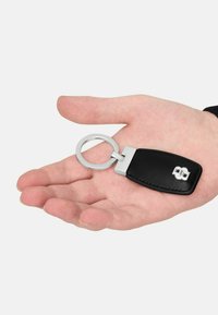 Czarny skórzany brelok z okrągłym srebrnym kółkiem i prostokątnym korpusem. Posiada wytłoczony wzór po jednej stronie. Gładka tekstura.