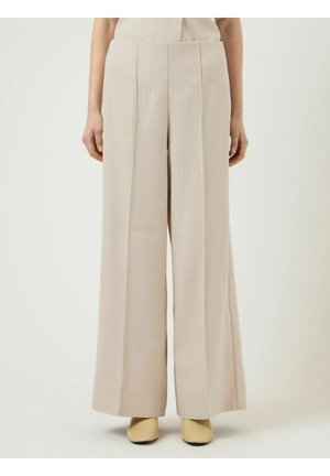 Pantalon beige taille haute à jambes larges porté avec un haut assorti et des chaussures à bouts pointus beige clair, debout devant un fond uni.