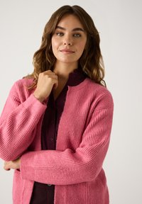 Rosa gerippter Cardigan aus weichem Material, mit langen Ärmeln und einem offenen Frontdesign. Über einer dunklen Bluse getragen.