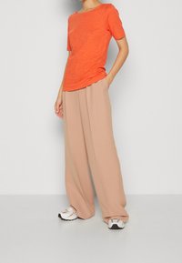 Top arancione a maniche corte con vestibilità rilassata, abbinato a pantaloni larghi color beige chiaro e sneakers bianche con dettagli grigi.