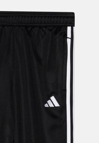 Melni sporta bikses ar gludu tekstūru, ar baltām sānu svītrām un sudraba Adidas logotipu apakšējā labajā kājā.