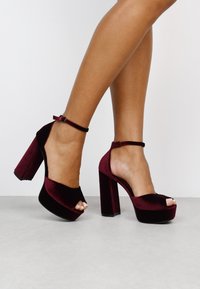 Sandalias de tacón alto de terciopelo burdeos con diseño de punta abierta, correa en el tobillo y tacones gruesos. Textura suave y acabado brillante.