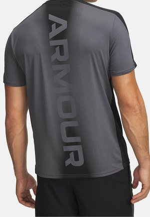Mann trägt ein dunkelgraues, kurzärmeliges Sportshirt mit dem Wort "ARMOUR" vertikal in großen Buchstaben auf dem Rücken gedruckt.