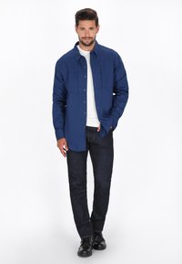 Camicia blu Navy a bottoni con due tasche frontali, indossata sopra una t-shirt bianca, abbinata a pantaloni scuri e scarpe nere. Tessuto morbido, vestibilità casual.