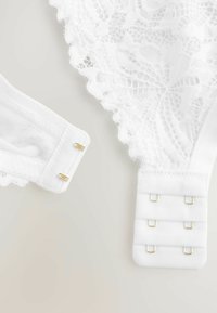 Filo del reggiseno in pizzo bianco con bordo ondulato, caratterizzato da un tessuto morbido e una leggera lucentezza. Include una chiusura a gancio e occhiello con hardware dorato.