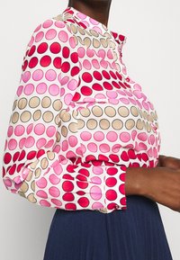 Blus med långa ärmar som har ett cirkulärt mönster i rosa, rött och beige på en vit bakgrund, med krage och knappslå.