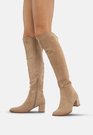 Beige suedine laarzen tot boven de knie met een blokhak, puntige neus en een rits aan de zijkant voor eenvoudig aan- en uittrekken. Gladde textuur met minimale accenten.