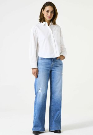 Weißes, kurz geschnittenes Hemd mit Knöpfen, Kragen und Tasche, kombiniert mit hellblauen, weit geschnittenen Jeans mit Used-Look. Schwarze Schuhe sichtbar.