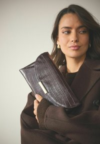 Dunkelbraune Clutch aus Kunst-Krokodilleder mit einer geschwungenen Klappe und goldfarbenem Verschluss. Weiche Textur, gehalten von einer Person in einem braunen Mantel.