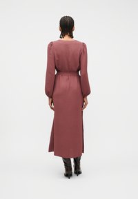 Γυναίκα με πλ εξούδες, φορεμένη με μακρύ μωβ φόρεμα με φουσκωτά μανίκια και μαύρες μπότες με ψηλό τακούνι, στεκόμενη με την πλάτη προς τα εμπρός σε λευκό φόντο.