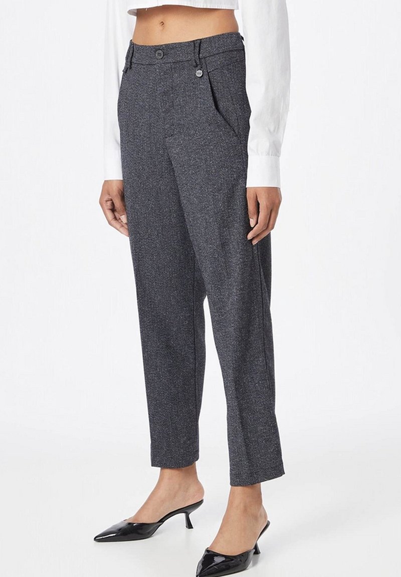 Nainen, joka käyttää korkevavyötäröisiä harmaita räätälöityjä housuja, joissa on etutaitokset, yhdistettynä valkoiseen pitkähihainen lyhyt puseroon ja mustiin matalakorkoisiin kenkiin.