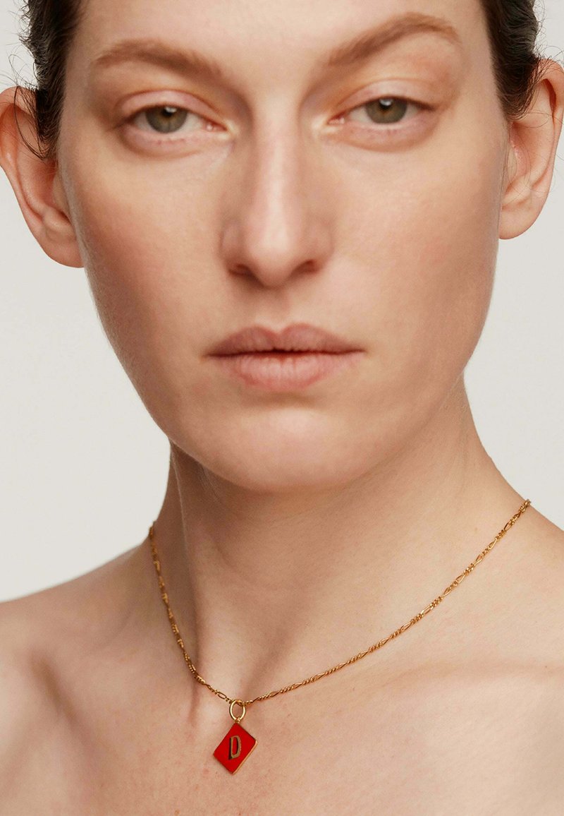 Collar de cadena torcida de oro con un colgante en forma de diamante rojo que presenta una "D" mayúscula en el centro. Textura suave y diseño minimalista.