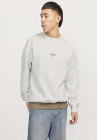 Light gray sweatshirt with dropped shoulders, ribbed cuffs, and a small black text logo. Worn over a brown layer and paired with blue jeans.