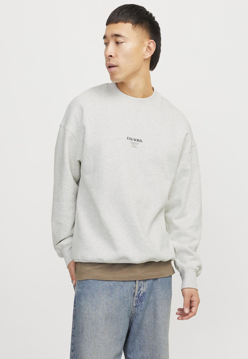 Light gray sweatshirt with dropped shoulders, ribbed cuffs, and a small black text logo. Worn over a brown layer and paired with blue jeans.