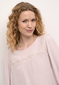Lys pink bluse med rund halsudskæring, der har dekorativt blondertrim og lange ærmer. Fremstillet af glat, let stof.
