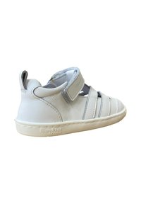 Zapato infantil de cuero blanco con correa de velcro ajustable, lados abiertos y suela de goma texturizada. Presenta sutiles acentos en gris y branding.