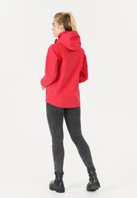 Röd vattentät jacka med huva, som har en stilren design, justerbara manschetter och en diskret logotyp på sidan. Bärs med mörka jeans och stövlar.