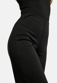 Detailný záber na čierne vysokopásové nohavice na telo, nosené s čiernym topom na jednoduchom svetlom pozadí.