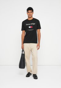 Musta puuvillane T-särk, millel on "Tommy Hilfiger" logo ja lipu graafika, kombinatsioonis beežide pükstega ja musta kotiga, jalas mustad kingad.