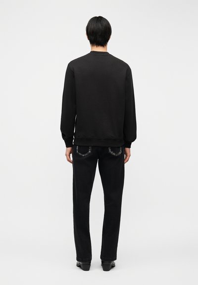 Personne debout, vue de dos, portant un sweat-shirt noir à manches longues, un jean noir avec des poches arrière cousues et des chaussures noires sur fond blanc.