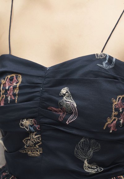 Gros plan sur une robe bleu foncé à fines bretelles ornée de motifs floraux et d'oiseaux brodés en fils rose, beige et bleu clair.