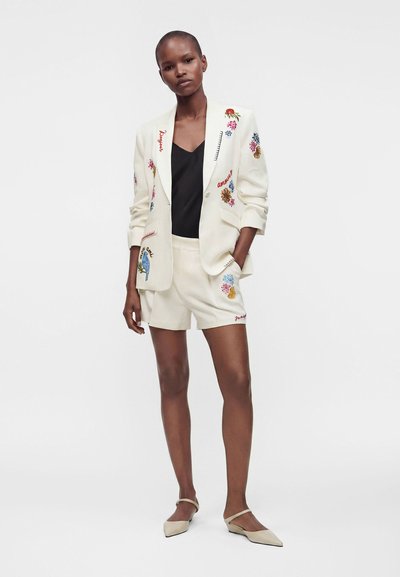 Femme debout en blazer blanc brodé et short assorti, top noir et ballerines crème sur fond blanc uni.
