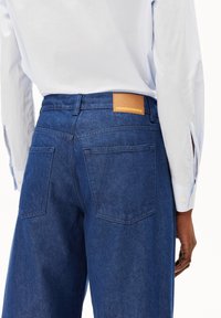 Dunkelblaue Denim-Jeans mit geradem Schnitt, ausgestattet mit einem beige Lederpatch, das "ARMEDANGELS" trägt, und zwei Gesäßtaschen. Ein weißes Hemd ist teilweise sichtbar.