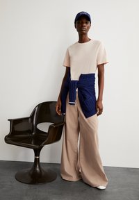 Beige T-shirt met korte mouwen, brede beige broek, marineblauwe gebreide trui bevestigd om de taille, marineblauw pet, zittend op een donkere kunststof stoel.