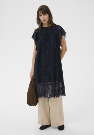 Kobieta w granatowej koronkowej sukience na beżowych szerokich spodniach, trzymająca brązową zamszową torbę, stojąca na jednolitym jasnym tle.