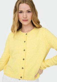 Gul cardigan laget av myk strikket materiale, med rund hals og seks mørke knapper nedover fronten. Lange ermer med ribbestrikkede mansjetter.