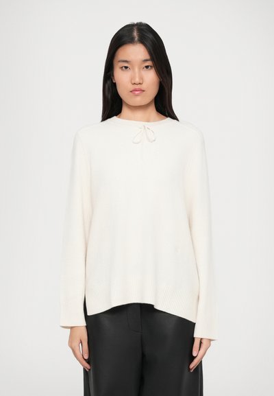 Pull léger crème en tissu doux, avec un col rond et un détail de nœud à l'avant, porté par-dessus un pantalon en cuir noir.