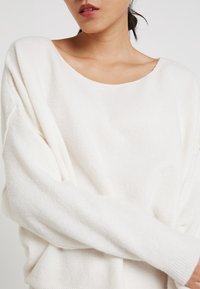 Vit oversized tröja med vid halsringning, långa ärmar och mjukt, texturerat tyg. Enkel design utan mönster eller anmärkningsvärda detaljer.