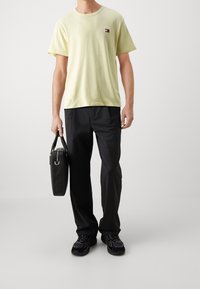 Light yellow cotton t-shirt with round neck and small logo, paired with black trousers and black sneakers, holding a black bag.