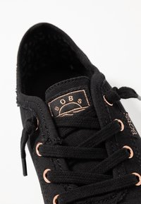 Czarny tenisówka na płótnie z okrągłym noskiem, miedzianymi przelotkami i fakturowaną czarną wkładką. Język buta zdobi logo w jasnym miedzianym kolorze.