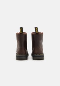 Botas de tobillo de cuero marrón con suelas de goma negra y costuras amarillas, que cuentan con tiradores de tela en la parte posterior para facilitar su uso.