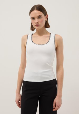 Femme aux cheveux bruns courts portant un haut blanc sans manches avec bordure noire et un pantalon noir, debout devant un fond clair uni.