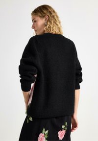 Černý, texturovaný pletený svetr s dlouhými, ohrnutými rukávy a širokým výstřihem; má jemně vyčesaný vzhled.