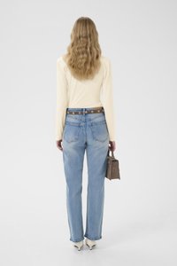 Lys beige langermet topp, høytlivs falmede blå jeans, brunt belte med metall detaljer, bærer en liten teksturert brun håndveske, hvite hæler.