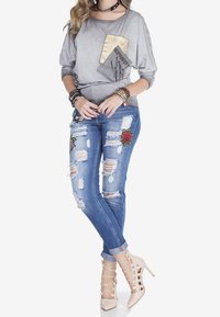 Camisa de manga larga gris con un bolsillo estampado, jeans azules desgastados con bordado floral y tacones altos color nude de tiras.