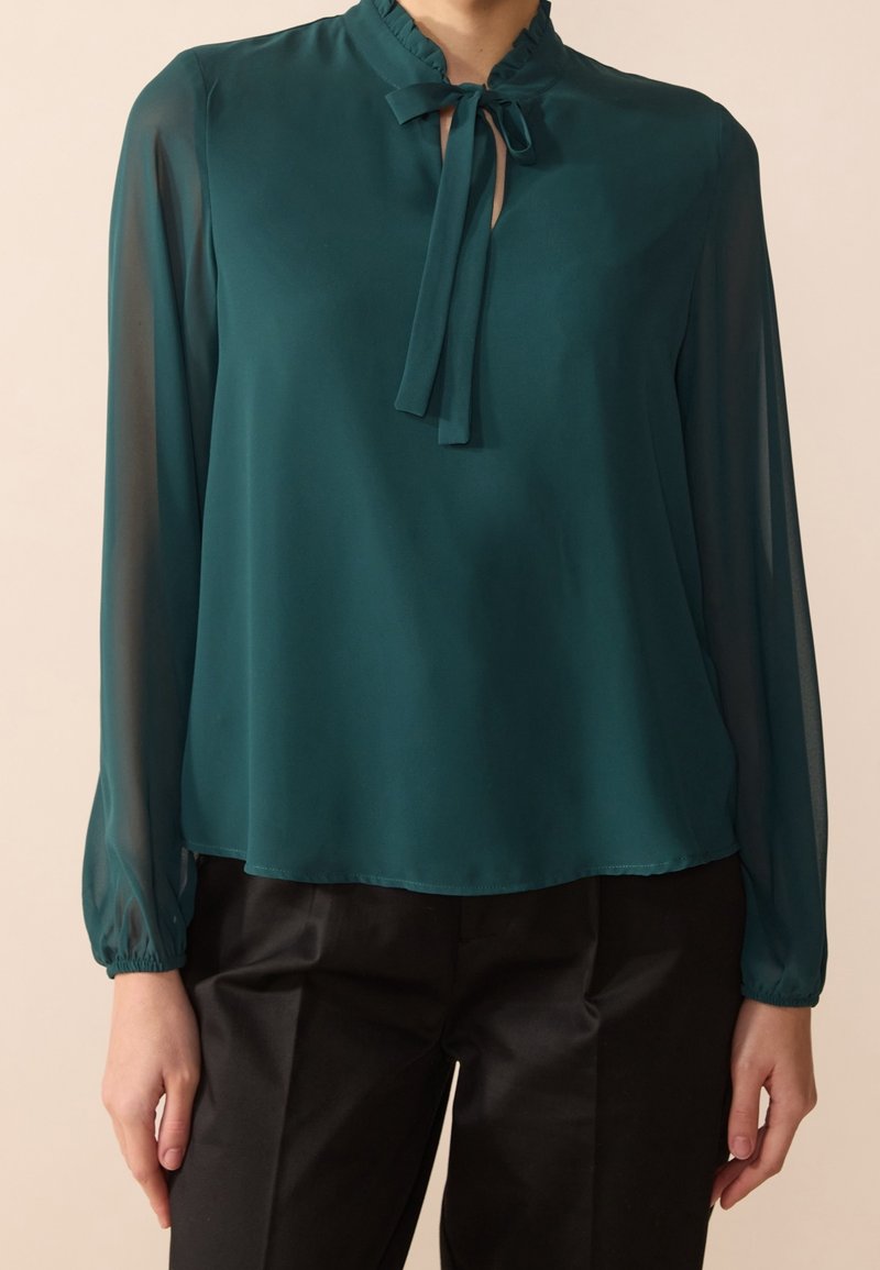Blusa trasparente turchese con scollo arricciato e chiusura con fiocco, maniche lunghe con polsini arricciati, indossata sopra pantaloni neri. Texture liscia.