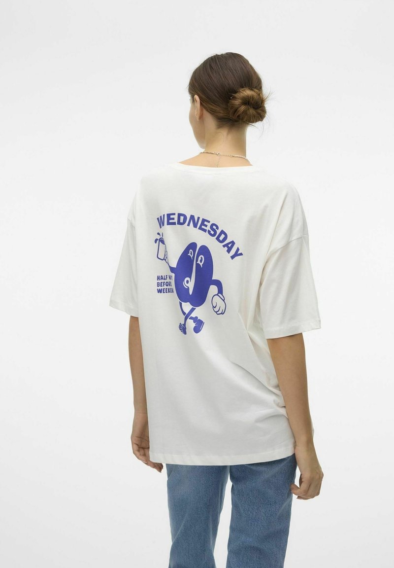 Femme avec un chignon porte un t-shirt blanc oversize avec un texte bleu "Wednesday" et un graphisme cartoon dans le dos, associé à un jean bleu.