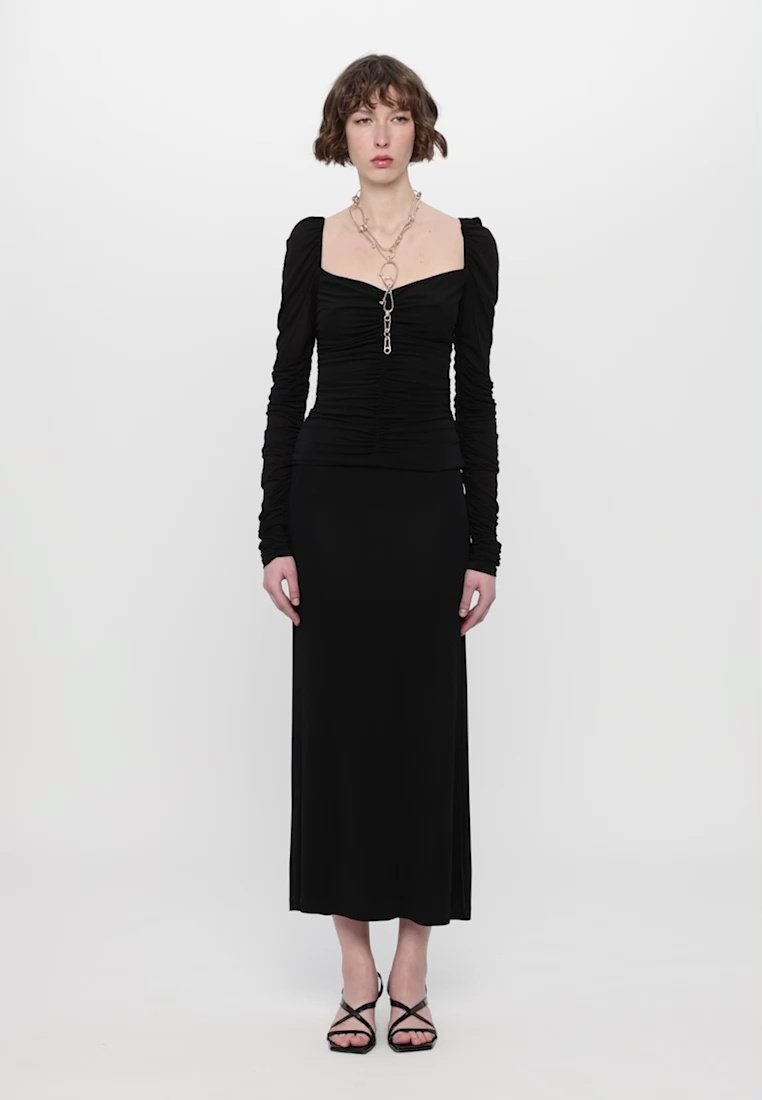 Haut noir ajusté à manches longues et détails froncés, associé à une longue jupe noire. Comprend un collier en chaîne avec un pièce maîtresse.