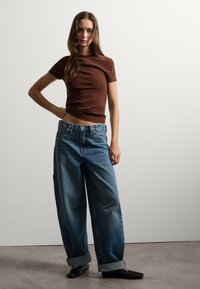 Bruine geribbelde top met korte mouwen, gecombineerd met oversized blauwe denim jeans. De jeans zijn iets vervaagd met opgerolde boorden, aangevuld met zwarte schoenen.